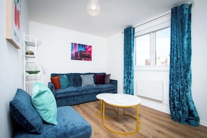 Ein modernes Wohnzimmer mit einem blauen Sofa, einem weißen Couchtisch, einem Fenster mit türkisfarbenen Vorhängen und einem Stadtbild an der Wand.