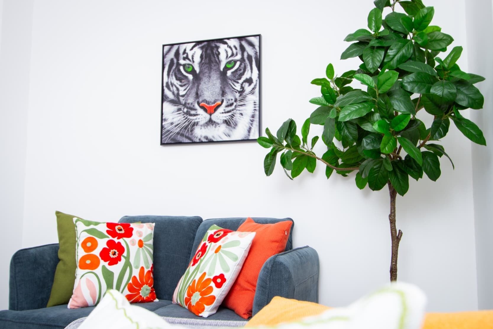 Ett vardagsrum med en soffa, kuddar, en inramad tavla med ett tigermotiv och en grön växt.