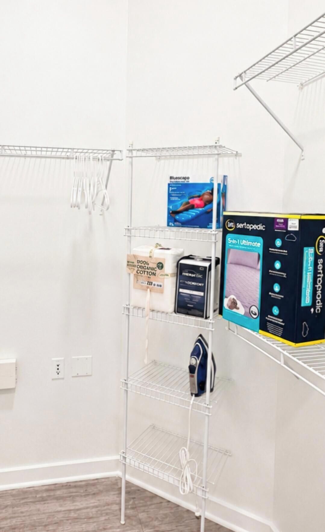 Um quarto branco com uma estante de metal que contém diversos objetos, incluindo uma caixa rotulada 'Sertapedic' e uma caixa azul com a imagem de uma pessoa.