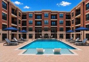 Una piscina rectangular con sillas de descanso y sombrillas, rodeada por un edificio de ladrillo con varios balcones.