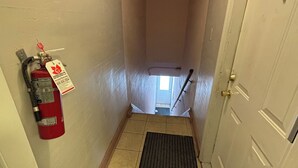 A narrow hallway with a fire extinguisher mounted on the wall, a doormat with floral patterns, and a door at the end.