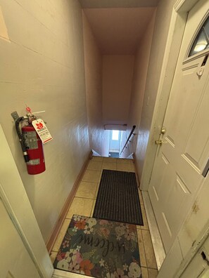 Ein schmaler Gang mit einem an der Wand befestigten Feuerlöscher, einer Fußmatte mit floralem Muster und einer Tür am Ende.