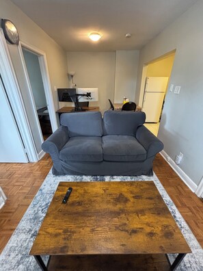 Ein Wohnzimmer mit grauem Sofa, einem Holztisch, einem Fernseher und einer Küche im Hintergrund.
