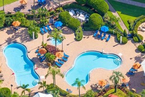 Vue aérienne d’un complexe hôtelier avec deux piscines, des chaises longues et des palmiers.