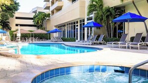 A swimming pool with lounge chairs and umbrellas, surrounded by a building with balconies. - 2BR Condo Near Fort Lauderdale Boat Show (FLIBS) (Pompano Beach)