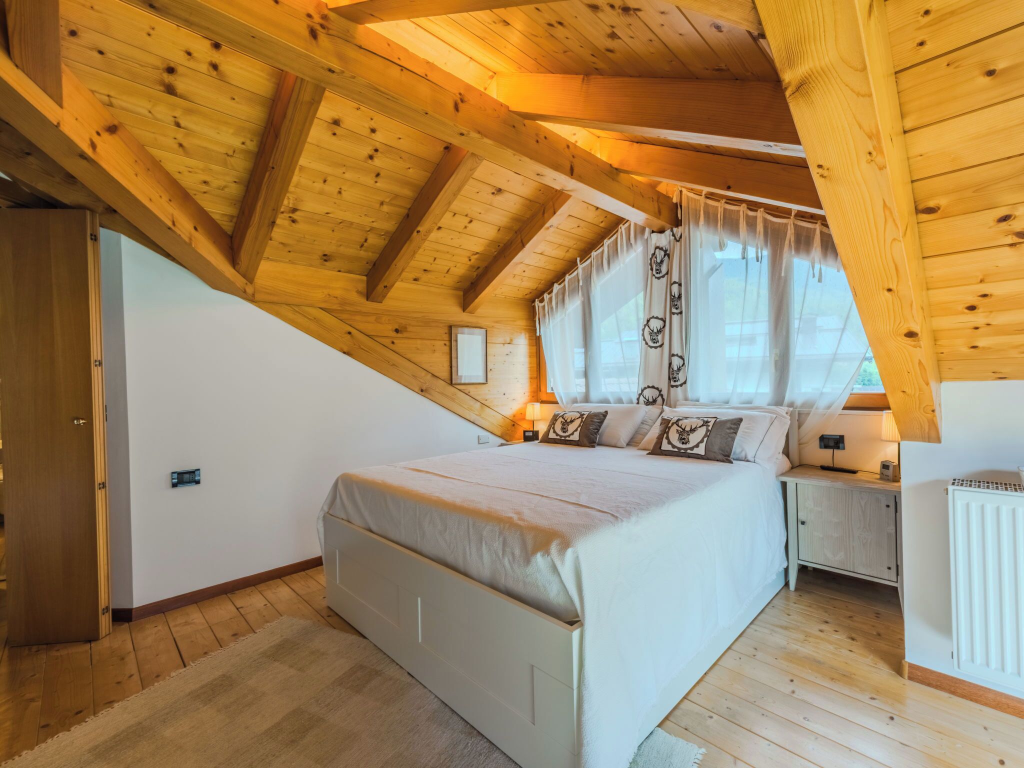 A bedroom with a sloped ceiling, a bed, bedside tables, a window with curtains, and a radiator.