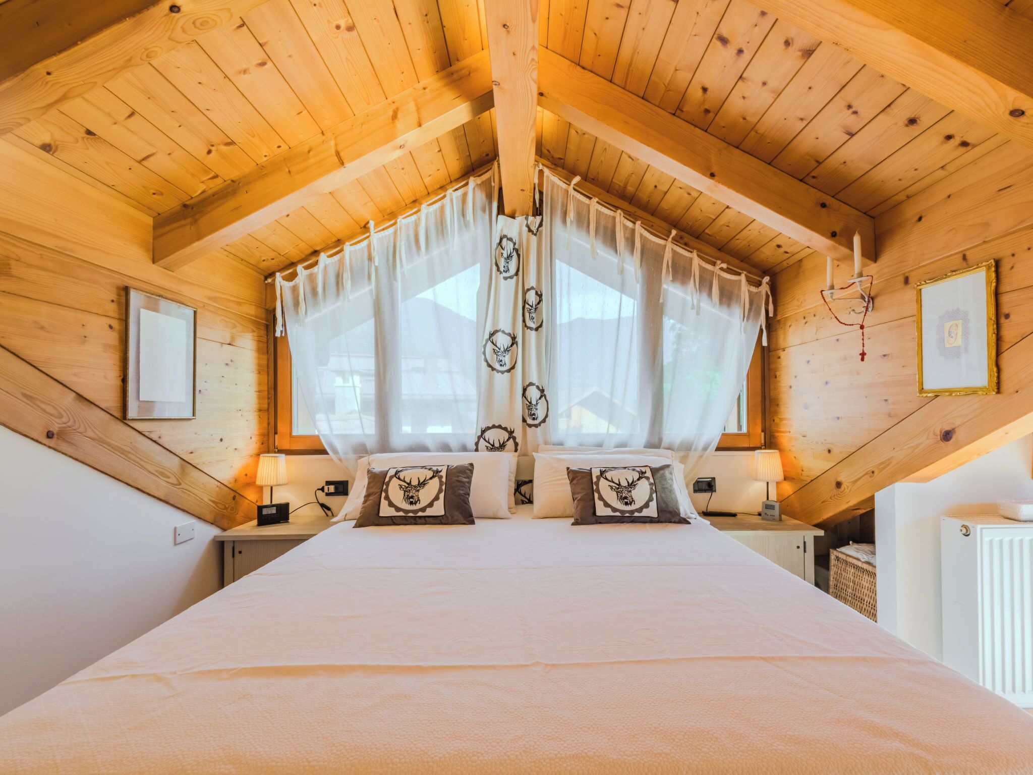 Un plafond en bois à pente douce, un lit recouvert d’une courtepointe blanche et agrémenté de coussins décoratifs, et une fenêtre ornée de rideaux légers.