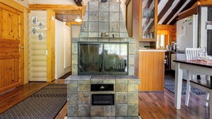 A stone fireplace with a glass door, wooden beams on the ceiling, and a kitchen area with a refrigerator and dining table. - Villa Succa (Kittilä)