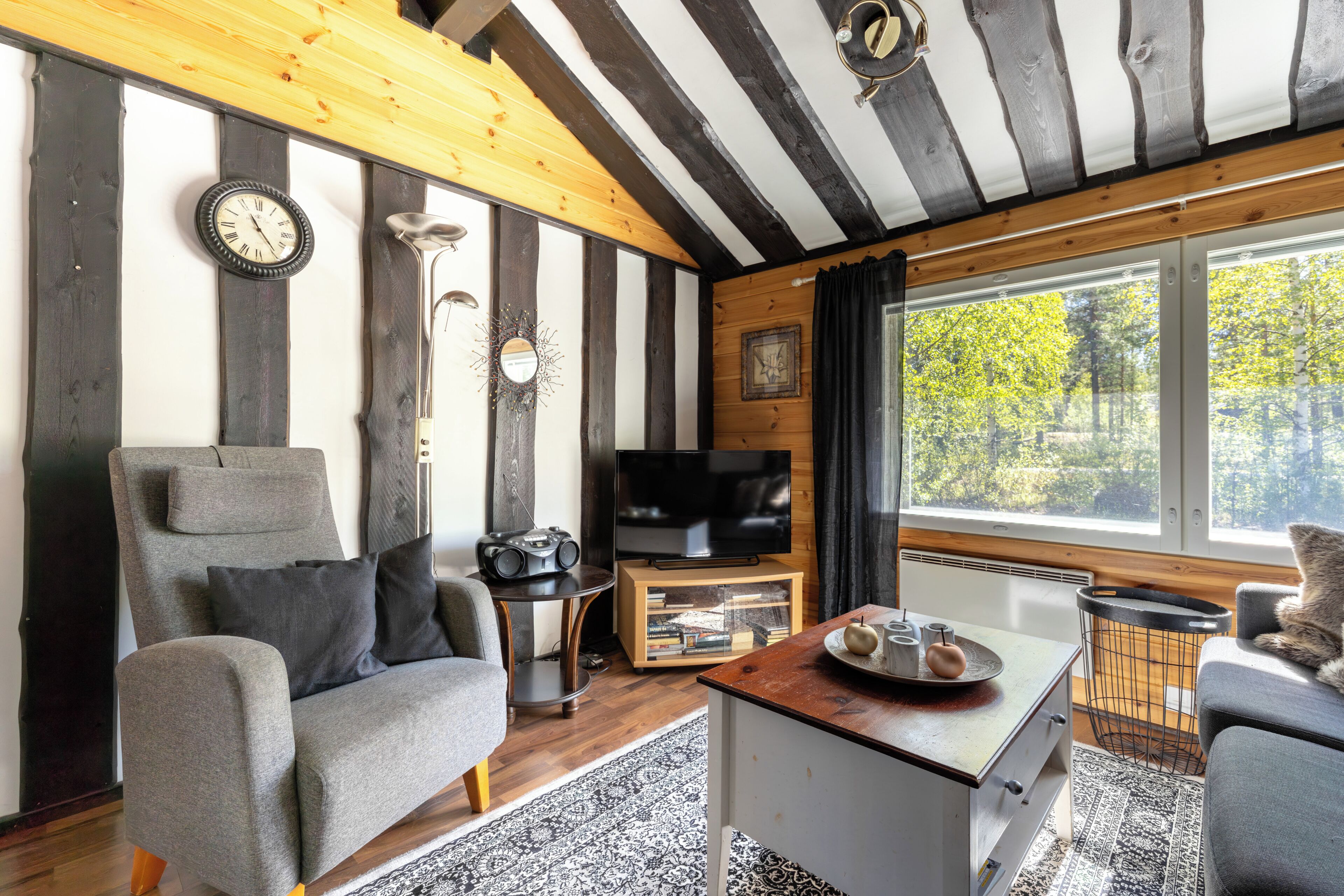 A room with a wooden ceiling, a television on a wooden stand, a grey sofa, a small table with a tray, and a window with a view of trees.