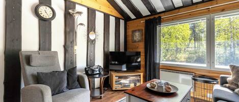 A room with a wooden ceiling, a television on a wooden stand, a grey sofa, a small table with a tray, and a window with a view of trees.