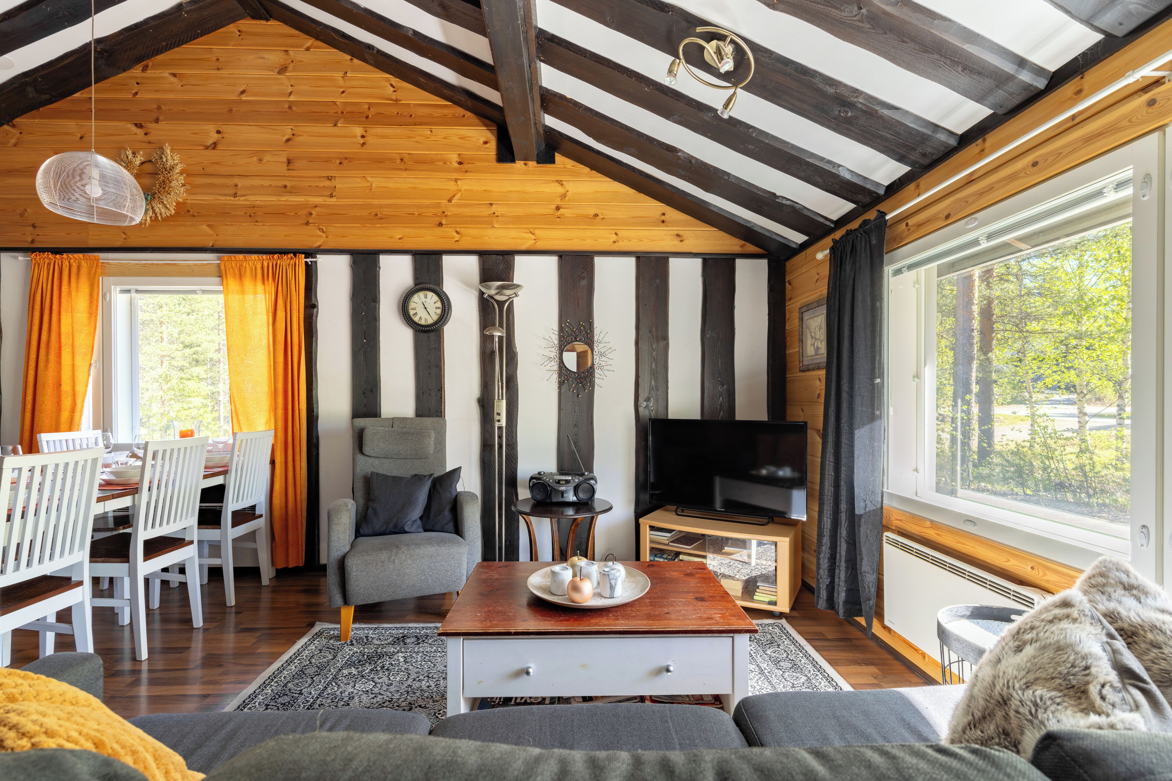 Un salon chaleureux avec un plafond en bois, un coin repas agrémenté de rideaux orange et une grande fenêtre donnant sur la forêt.