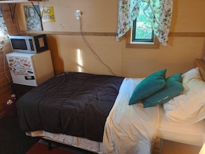 A small, compact room with a bed, a microwave, a refrigerator, and a window with floral curtains. - COZY PRIVATE TREE HOUSE (Saint John)