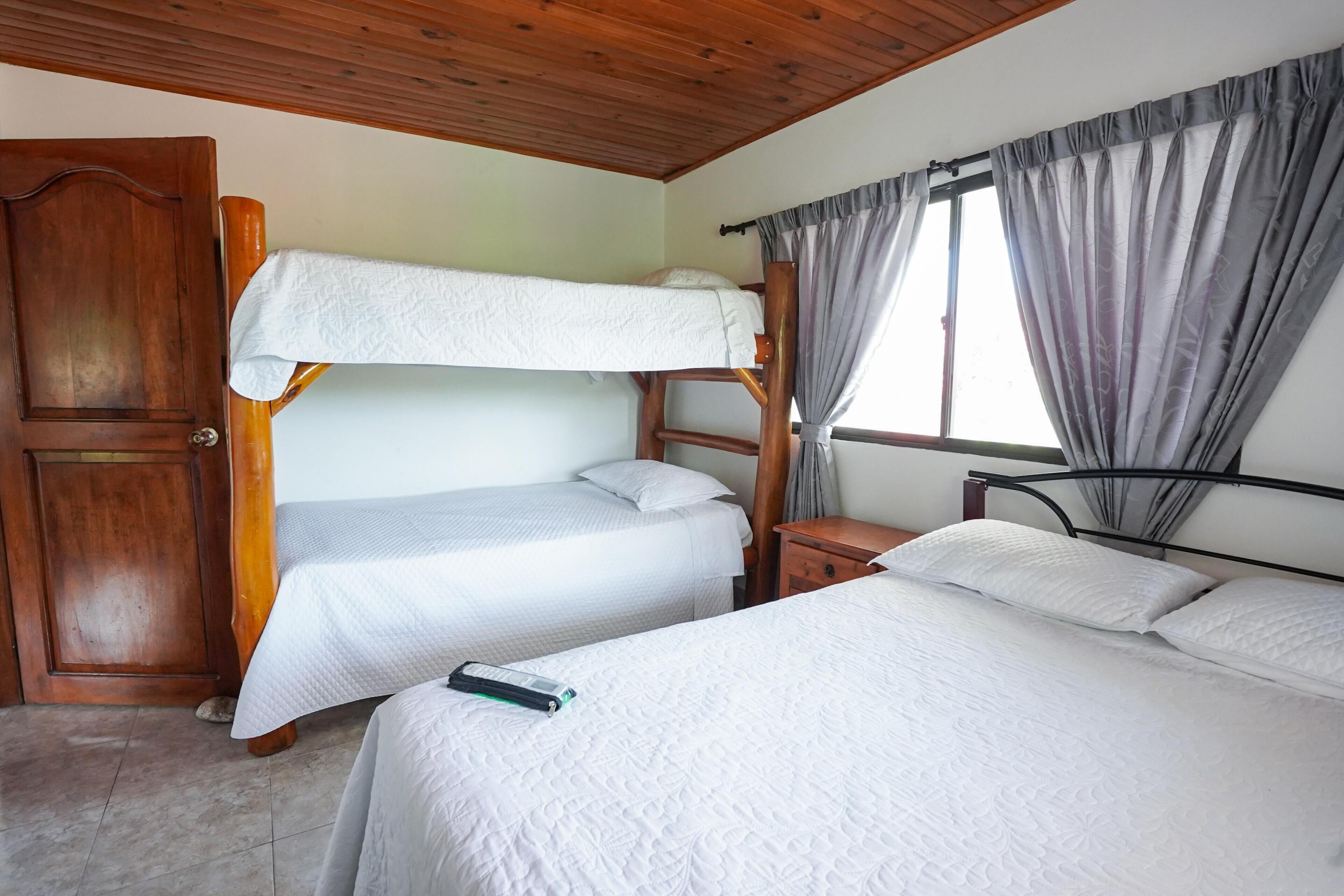 A room with bunk beds, a wooden door, a window with curtains, and a bedside table.