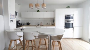Eine moderne Küche mit weißen Schränken, einer zentralen Kochinsel mit Hockern, hängenden Korbtropfenlampen und eingebauten Backöfen.