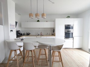 Uma cozinha moderna com armários brancos, uma ilha central com banquetas, luminárias pendentes de vime e fornos embutidos.
