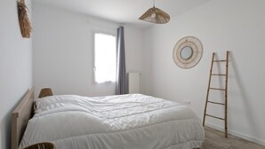 Ein Schlafzimmer mit Bett, Fenster, Pendelleuchte, Wandspiegel und einer Holzleiter.