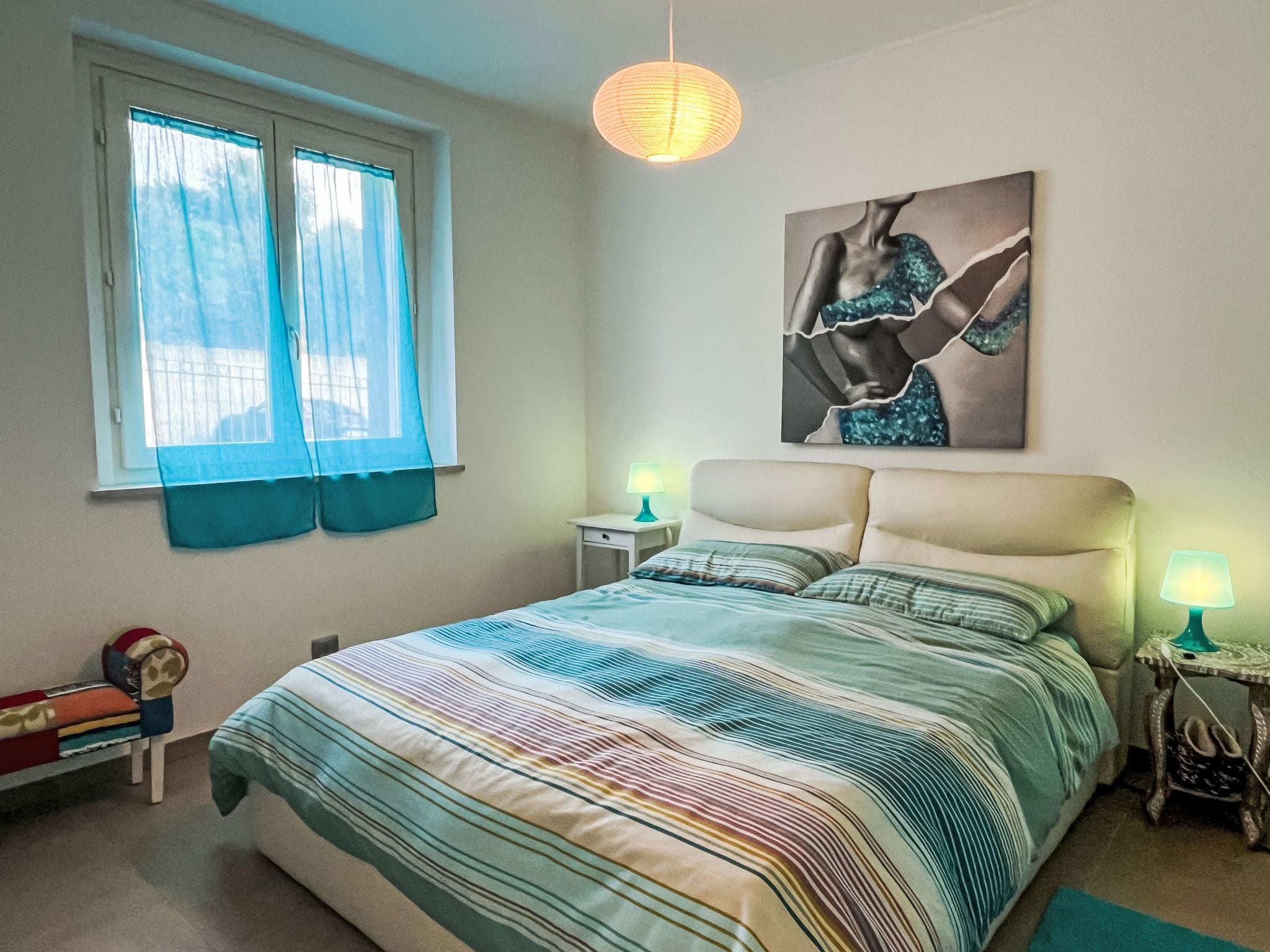 Quarto com cama, duas mesas de cabeceira com luminárias, janela com cortinas e uma obra de arte pendurada na parede.