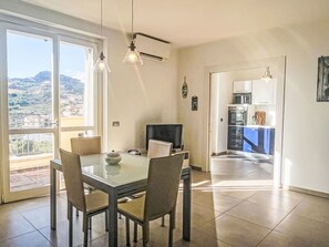 Un comedor con mesa de vidrio, sillas y lámparas colgantes. Se ve una cocina y un balcón con vistas a la ciudad.