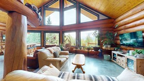 A log cabin interior with a sofa, armchair, and dining table, large windows, and a television. - Okanagan - Alpine Chalet (Christina Lake)