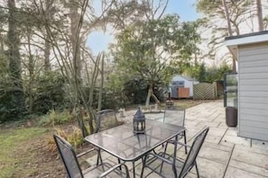 A patio with a table and chairs, a lantern, and a small shed in a wooded area. - Luxury6BedHouse-HotTub-GamesRoom (Poole)