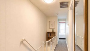 Un pasillo estrecho con barandilla blanca, una estantería y una puerta que da a otra habitación.