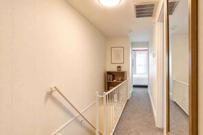 Un pasillo estrecho con barandilla blanca, una estantería y una puerta que da a otra habitación.
