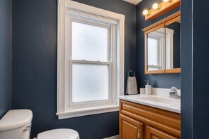 A bathroom with a white toilet, a window, a mirror, and a sink with wooden cabinets. - Fifa Stay Walk Manayunk River 4 Beds Sleeps 6 (Philadelphia)