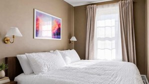 Una cama bien hecha con ropa blanca, un cuadro en la pared y una ventana con cortinas.