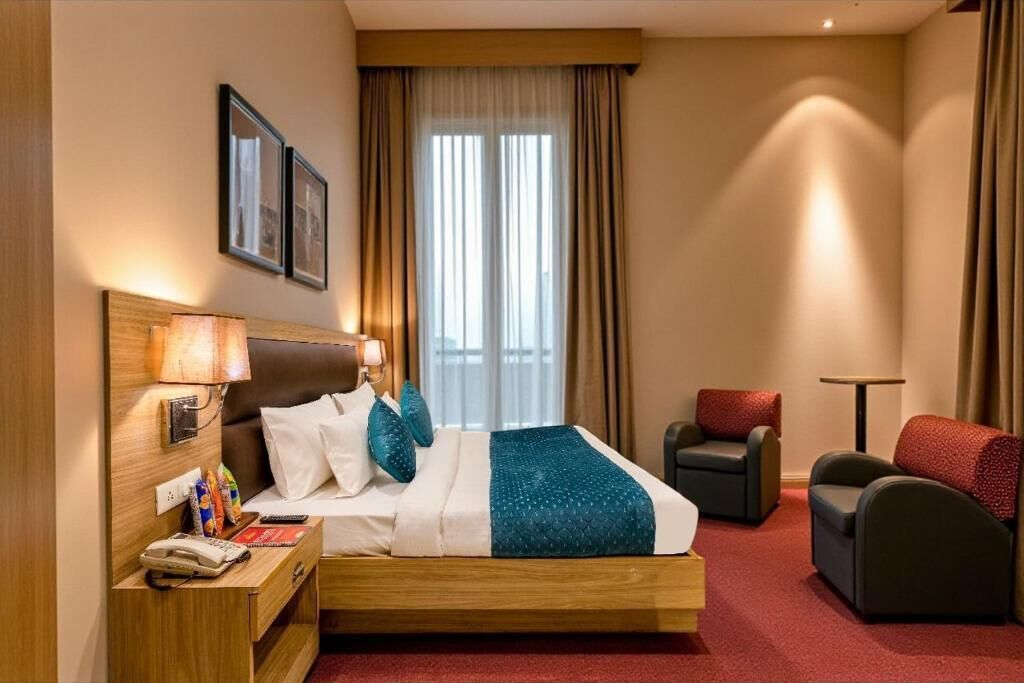 A hotel room with a bed, bedside table, lamp, chair, and a view of the outside through the window.