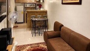 Un espace de vie compact comprenant un canapé marron, une table à manger avec des chaises, un coin cuisine avec des placards et des appareils électroménagers, et un ventilateur.