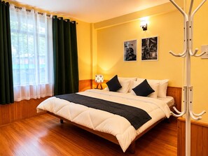 A bedroom with a bed, wooden floor, curtains, wall-mounted lamp, and two framed pictures. - HS GROUP OF HOTEL (Manali)