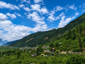 A mountainous landscape with a village nestled among the hills, featuring houses with colorful roofs and dense green vegetation. - HS GROUP OF HOTEL (Manali)