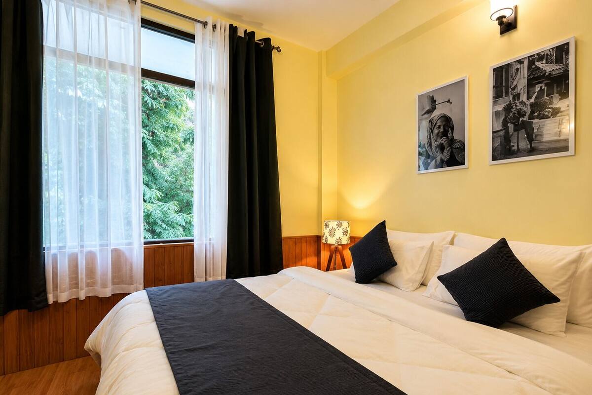 Habitación de hotel con cama, ventana con cortinas, lámpara y dos cuadros en la pared.