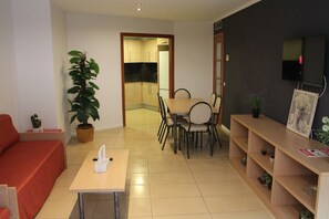 Un salon comprenant un canapé, une table à manger avec des chaises, un téléviseur fixé au mur et une plante en pot.