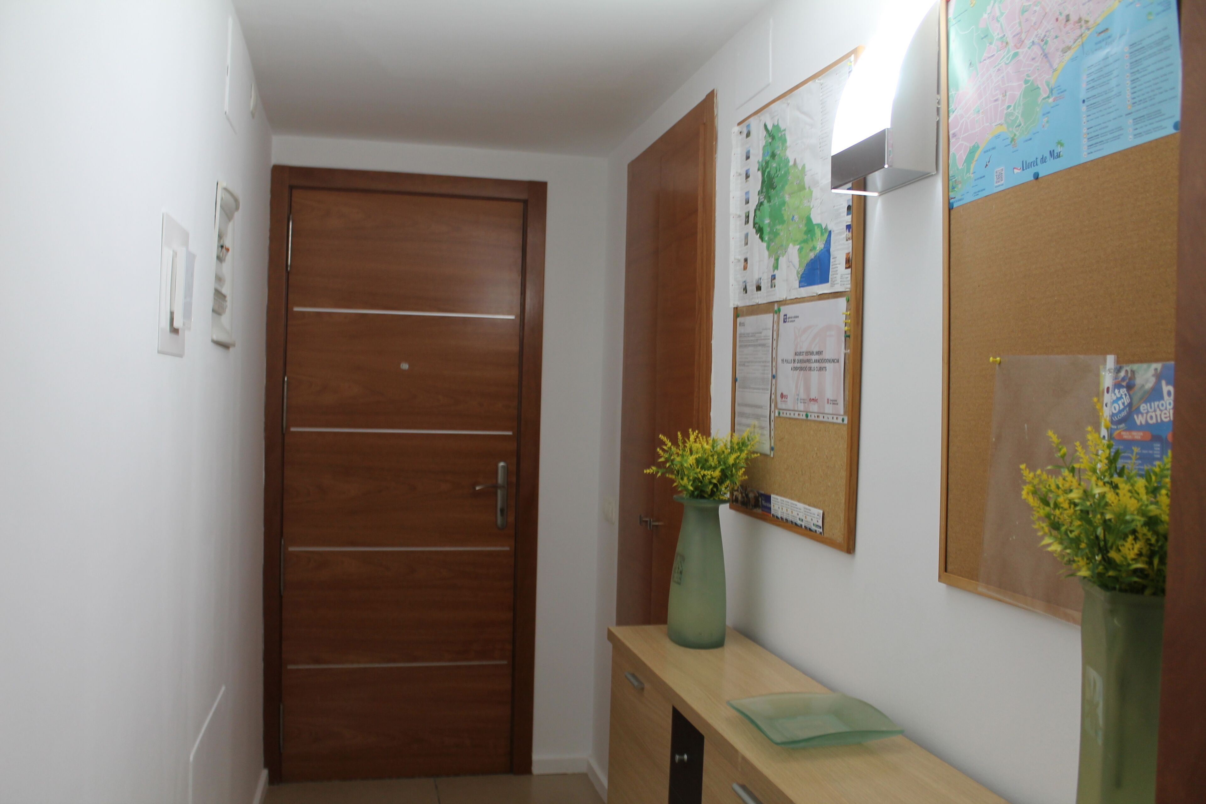 Un couloir avec une porte en bois, un tableau d’affichage orné de cartes et un petit bureau sur lequel se trouve un vase.