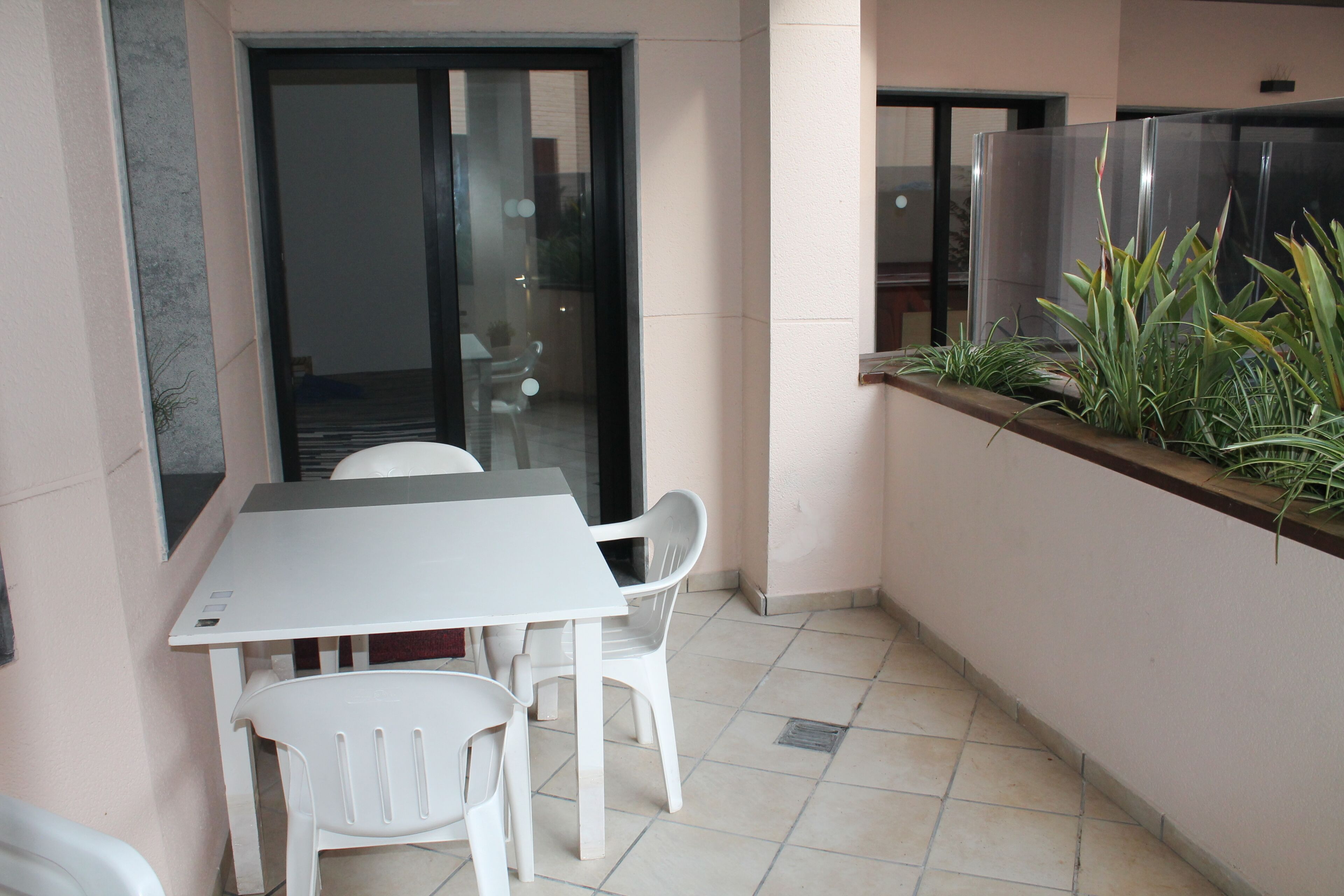 Un petit balcon avec une table et des chaises blanches, une porte vitrée donnant sur un espace intérieur et un jardinier contenant des plantes vertes.