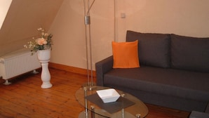 A living room with a glass coffee table, a grey sofa with an orange pillow, a tall floor lamp, and a radiator. - Maria Apartment 2 – Stay in the Heart of Wernigerode (Wernigerode)