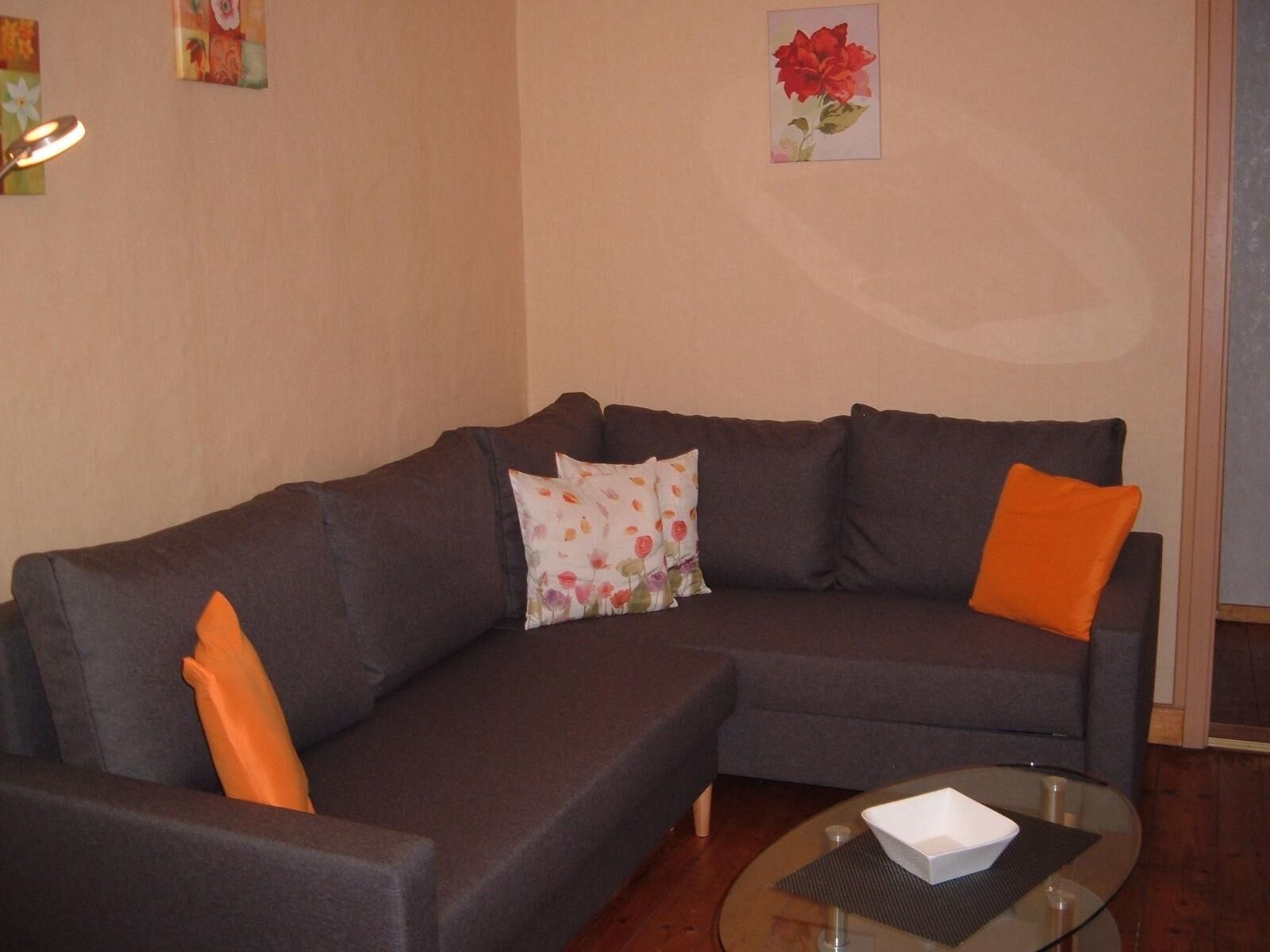 A living room with a dark-colored sofa, two orange cushions, a glass coffee table with a white object, and a painting of a red flower on the wall.