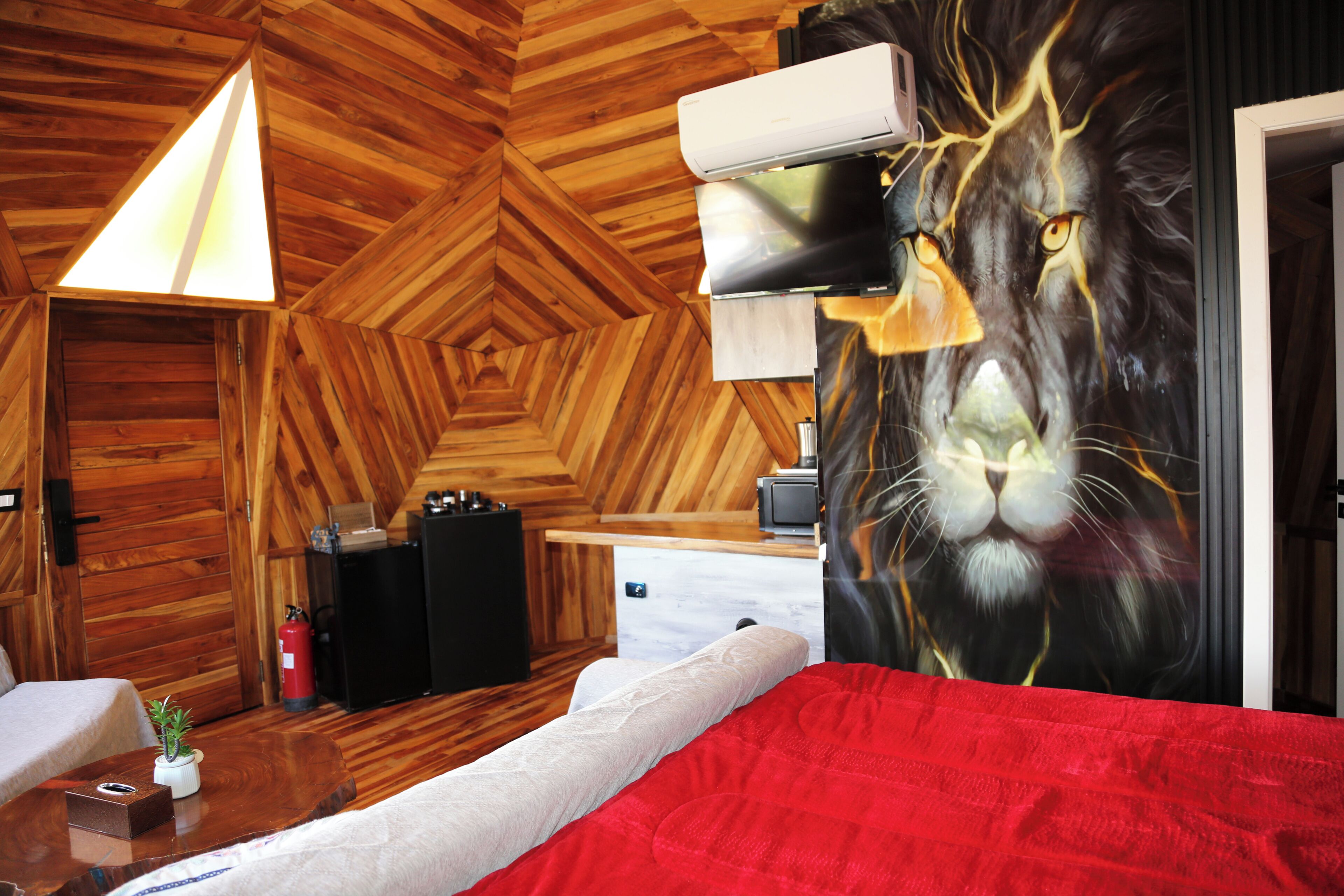 A room with a wooden herringbone floor, a bed with a red blanket, a wall-mounted air conditioner, a small table with a plant, and a painting of a lion's face.