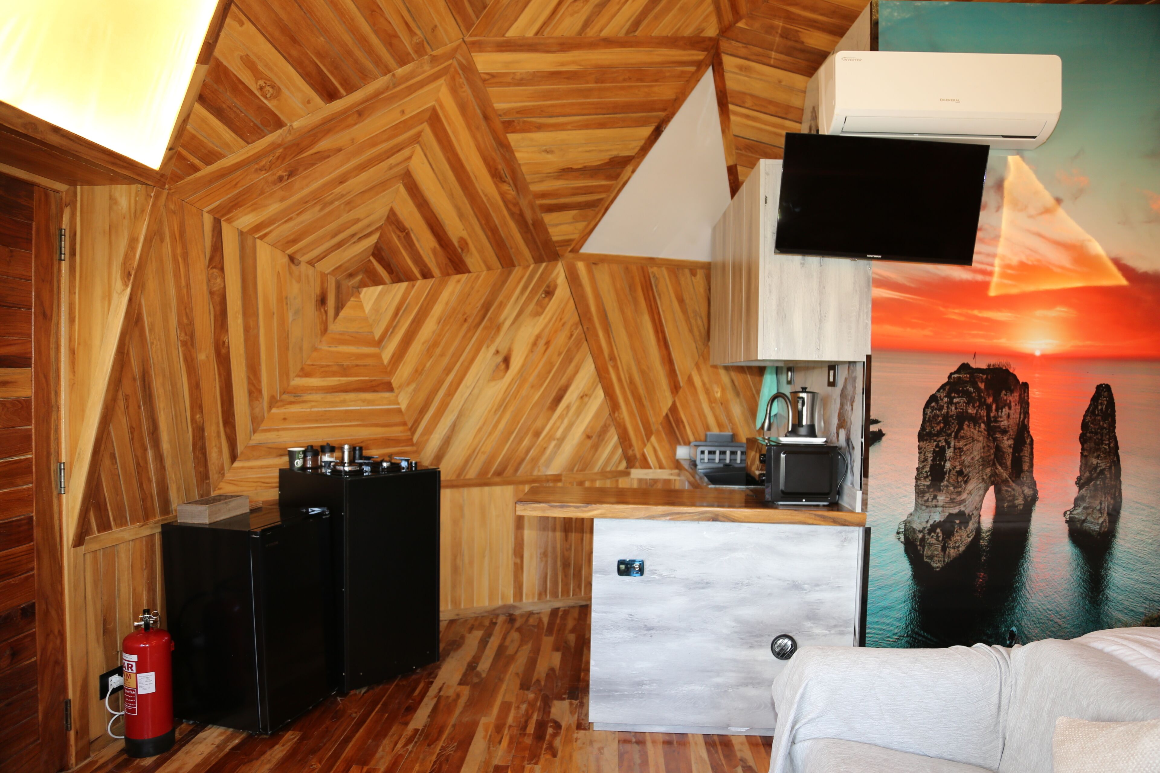 A room with a wooden herringbone ceiling, a flat-screen TV, a refrigerator, a fire extinguisher, and a painting of a sunset over rocks.