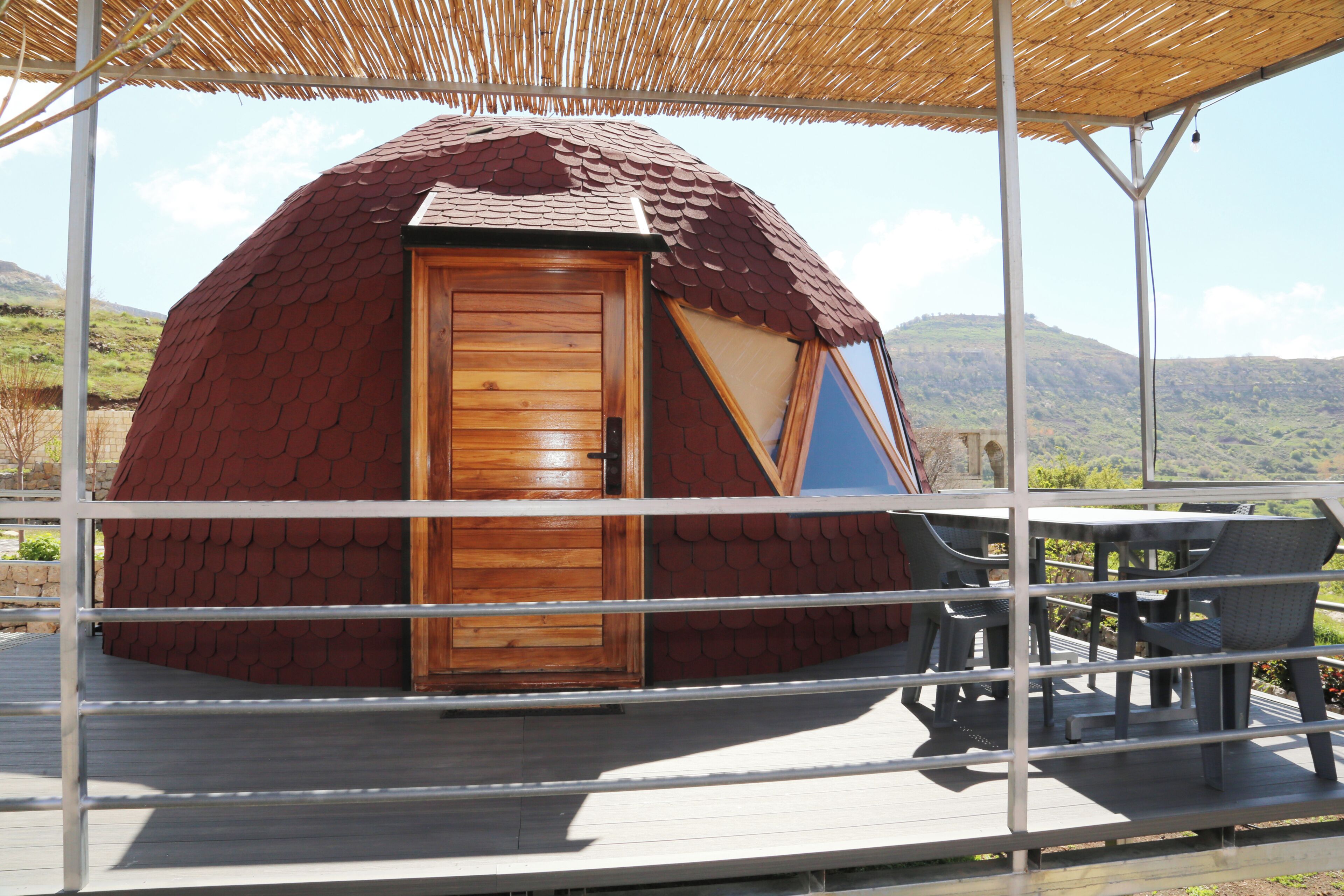 Una estructura de domo geodésico con puerta de madera, un área de estar cubierta al aire libre y vistas de un paisaje montañoso.