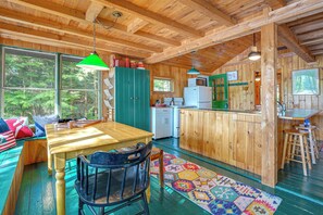 L’intérieur d’un chalet en bois comprend une table à manger, des chaises, un coin cuisine et une grande fenêtre donnant sur un paysage verdoyant.
