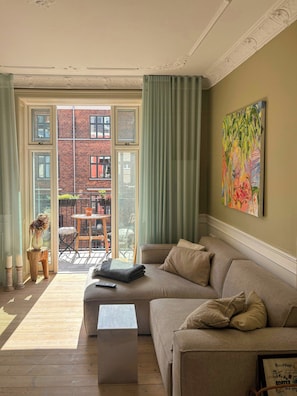 Ein Wohnzimmer mit Sofa, Couchtisch, einem Gemälde und Blick auf ein rot backsteins Gebäude durch eine Glastür.
