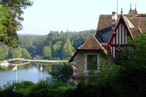 A traditional building with a stone facade, a prominent window, and a view of a lake with a bridge in the background.