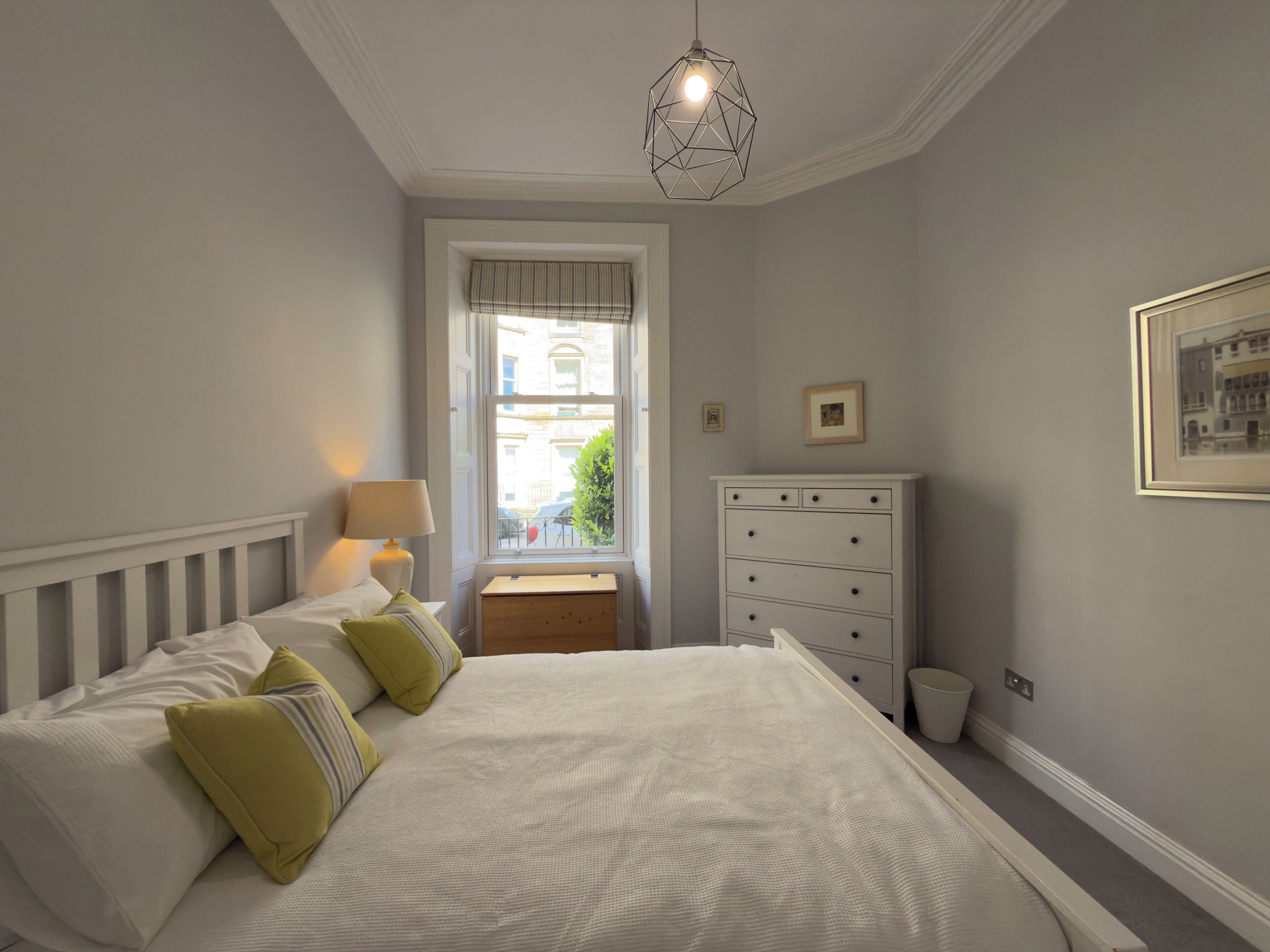 A bedroom with a bed, bedside lamp, dresser, window, and a hanging light fixture.