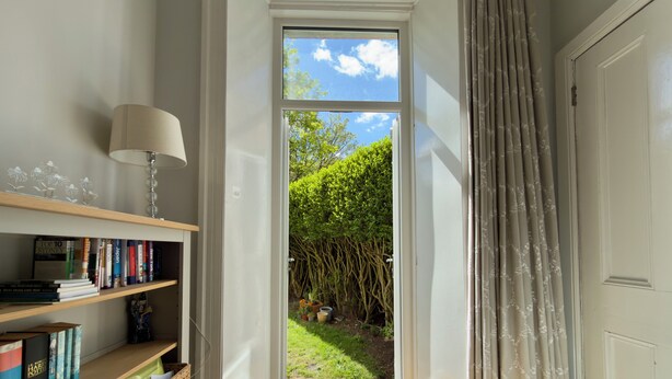 Ein Zimmer mit Bücherregal, Lampe, einer zur Gartenführenden Tür und einem mit Vorhängen versehenen Fenster.