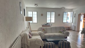 Ein geräumiges Wohnzimmer mit zwei weißen Sofas, einem Couchtisch und einer Stehlampe. Große Fenster sorgen für natürliches Licht, und eine Heizung ist an der Wand sichtbar.