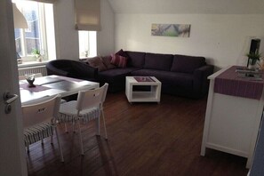 Ein Wohnzimmer mit einem dunkelgrauen Sofa, einem weißen Couchtisch, einem Esstisch mit Stühlen und einem Fenster mit Jalousien.