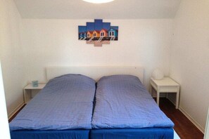 Ein kleines Zimmer mit einem Einzelbett, einem weißen Kopfteil, einer blauen Bettdecke und einem an der Wand befestigten Kunstwerk mit Häusern.