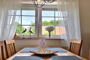 A dining area with a table, chairs, a woven centerpiece, a vase with flowers, and two bird sculptures on the windowsill. - What luck! - with a fence (Neßmersiel, Ostfriesland, Nordsee)